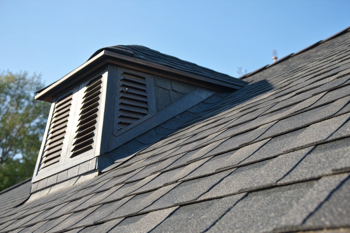 A louvered roof vent on a dark gray shingle roof against a blue sky